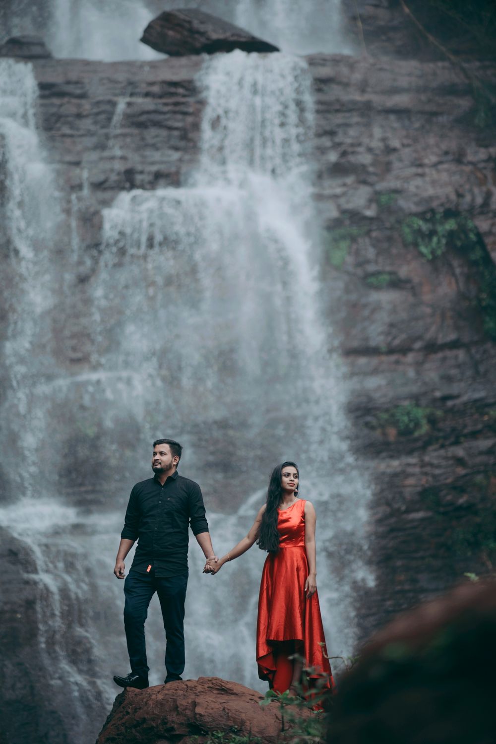 Couple holding hands in front of a majestic waterfall, woman in an elegant red gown.