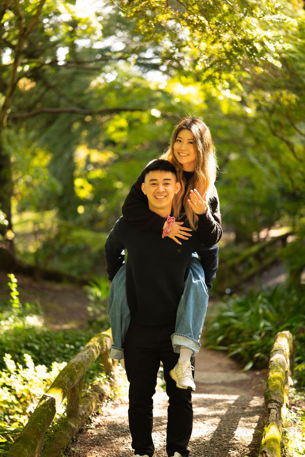 A smiling young man gives a happy young woman a piggyback ride on a sunlit garden path surrounded by lush green trees.