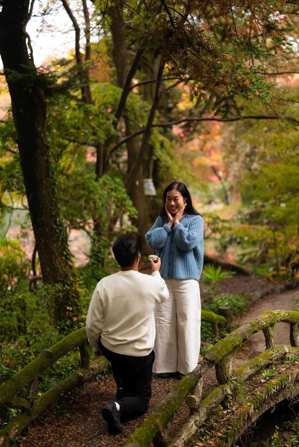 Man kneeling on garden path proposing to surprised woman in blue sweater among autumn trees.