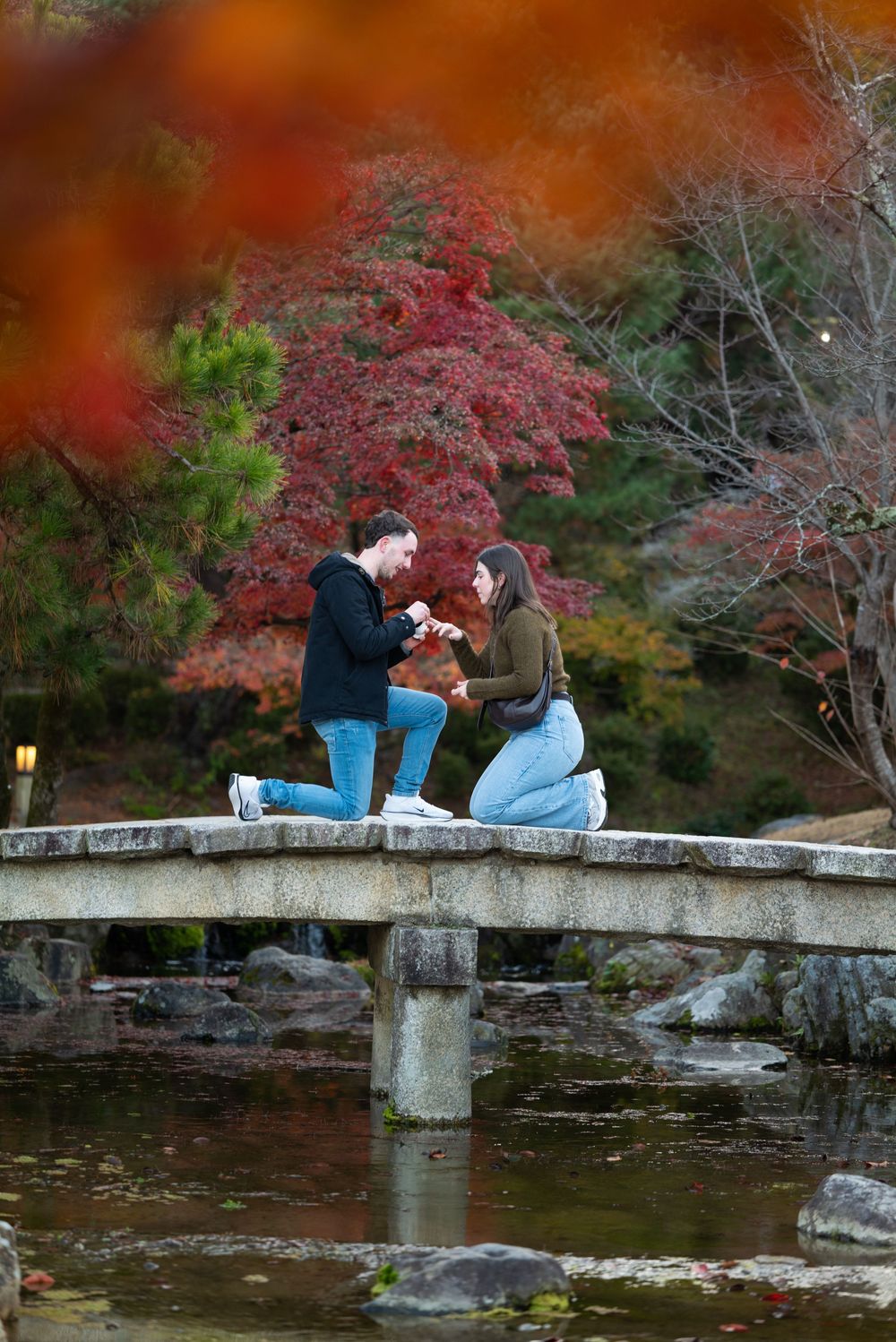 Couple sharing a romantic proposal moment on a stone bridge surrounded by vibrant autumn foliage in a Japanese garden.
