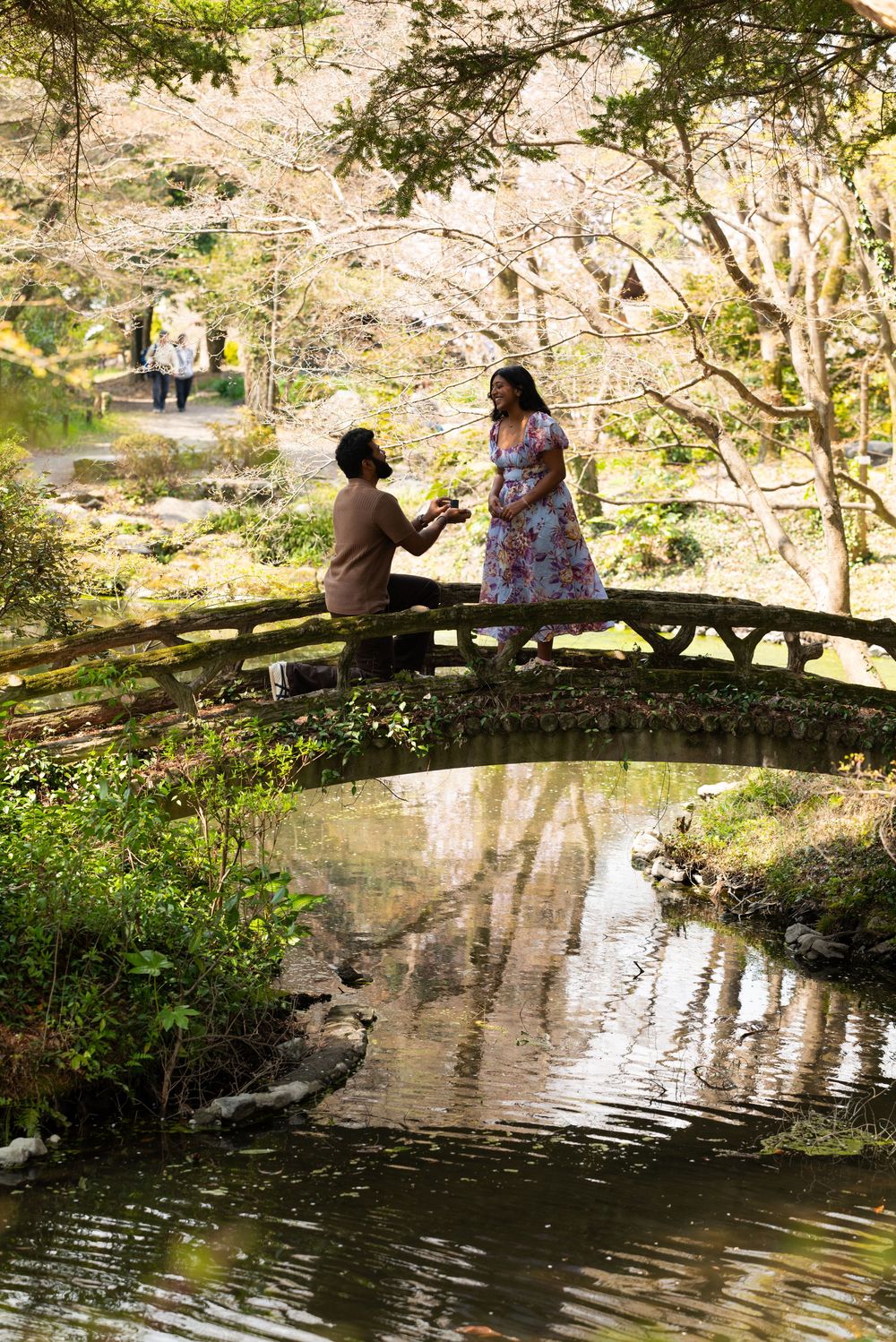 Couple sharing a romantic marriage proposal on a mossy stone bridge over a tranquil pond in a Japanese garden.