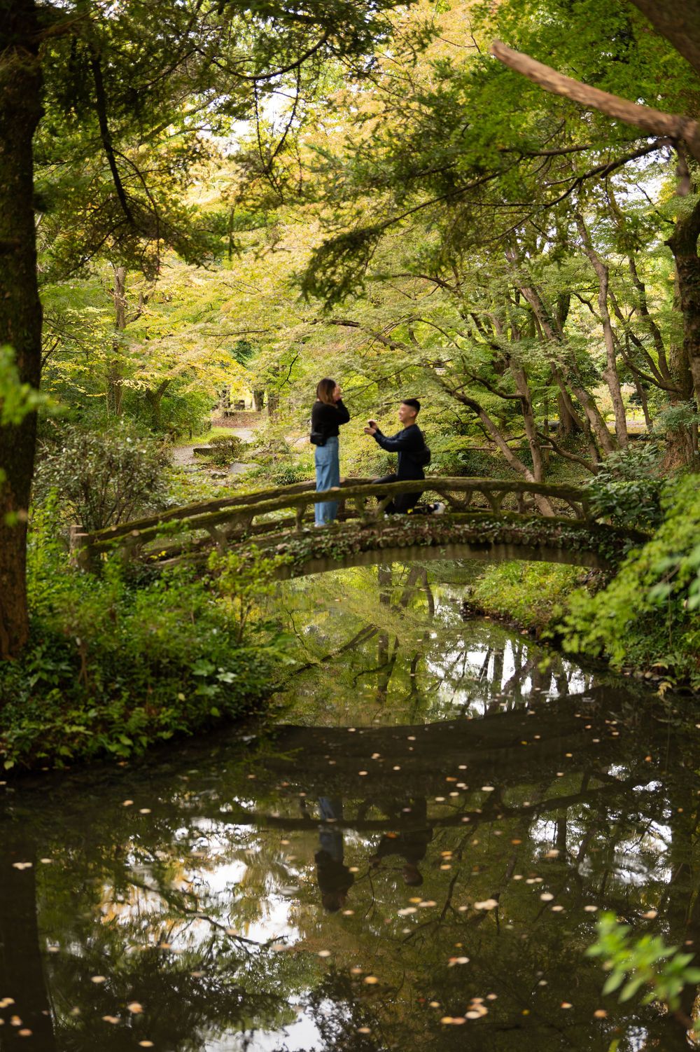 Two people stand on an arched wooden bridge reflected in a calm pond within a lush Japanese garden.