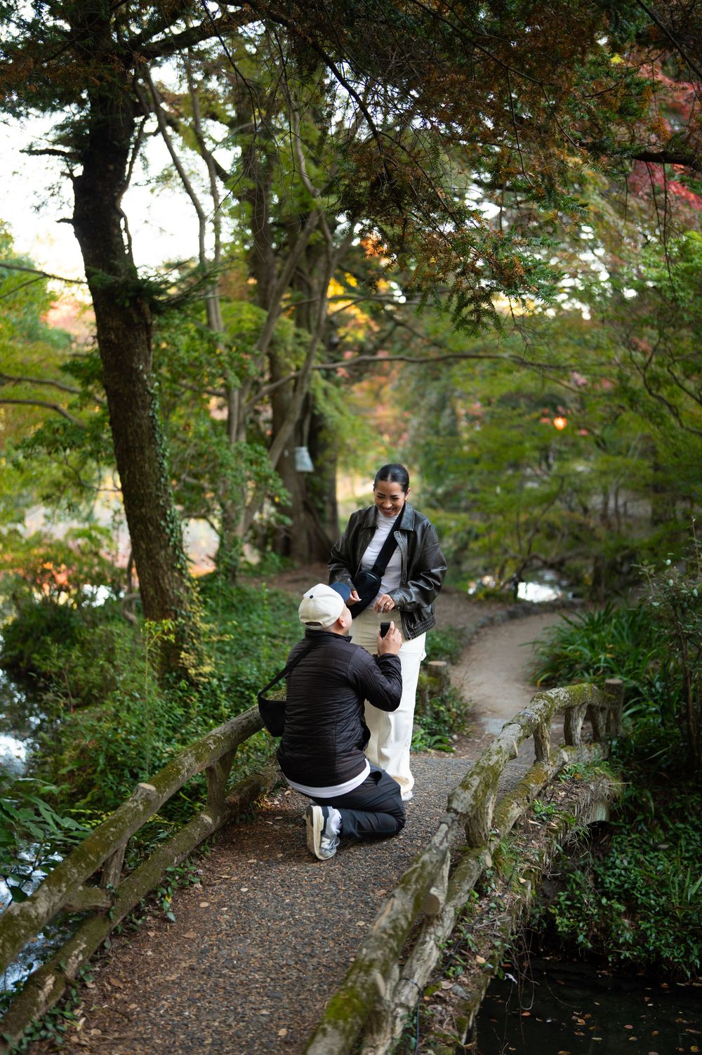 A person kneeling to propose to their partner on a scenic garden path surrounded by lush green and autumn-colored trees.