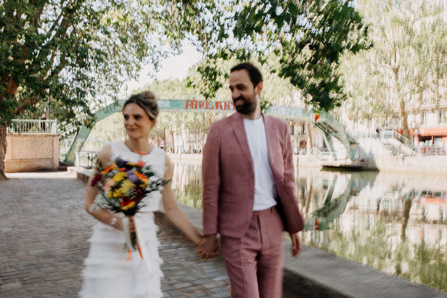 Bride and groom walk hand in hand along a scenic canal path, bride holding colorful bouquet, groom in pink suit.