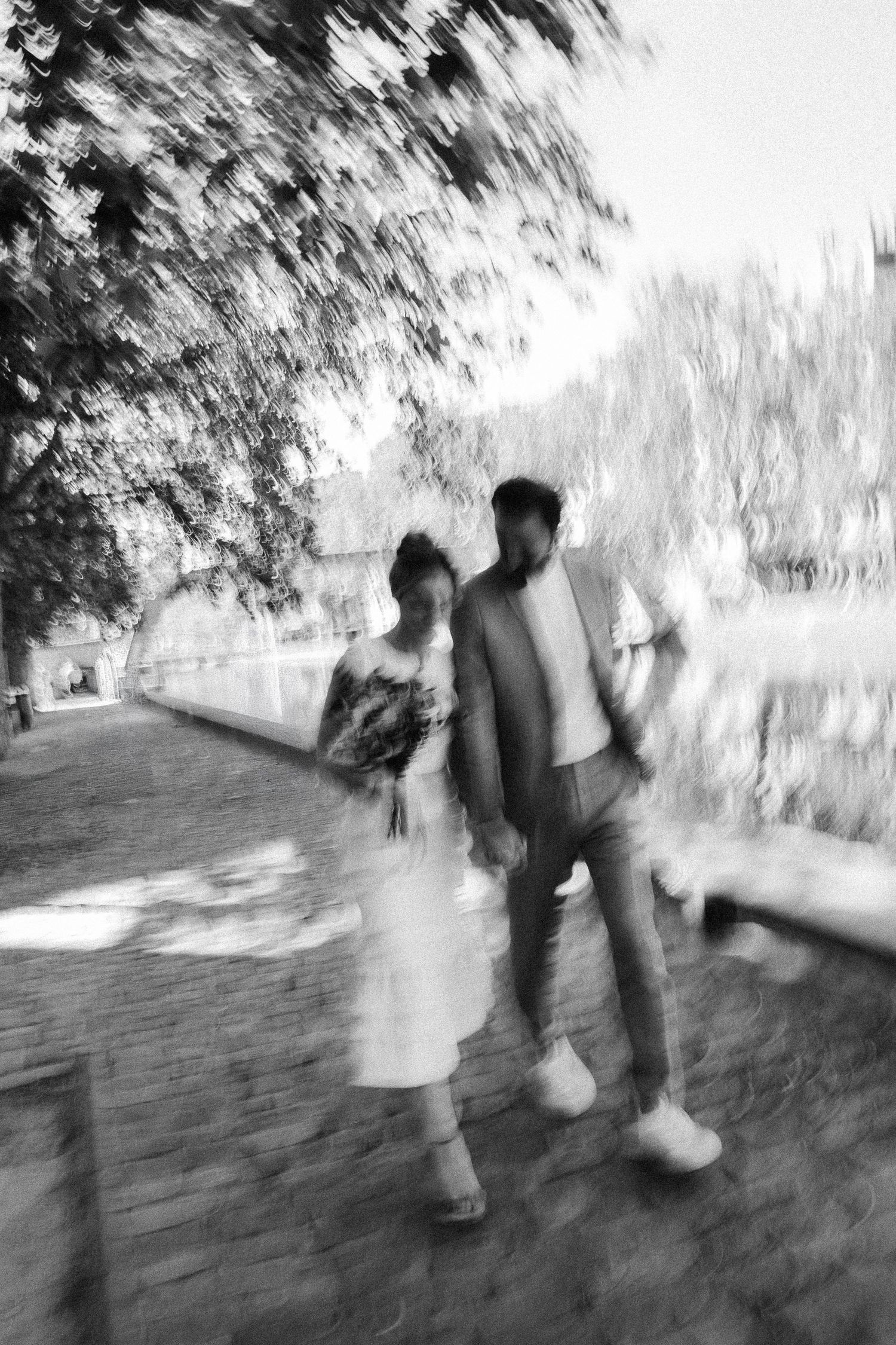Artistic blurred black and white photo of bride and groom walking along a tree-lined canal path on wedding day.