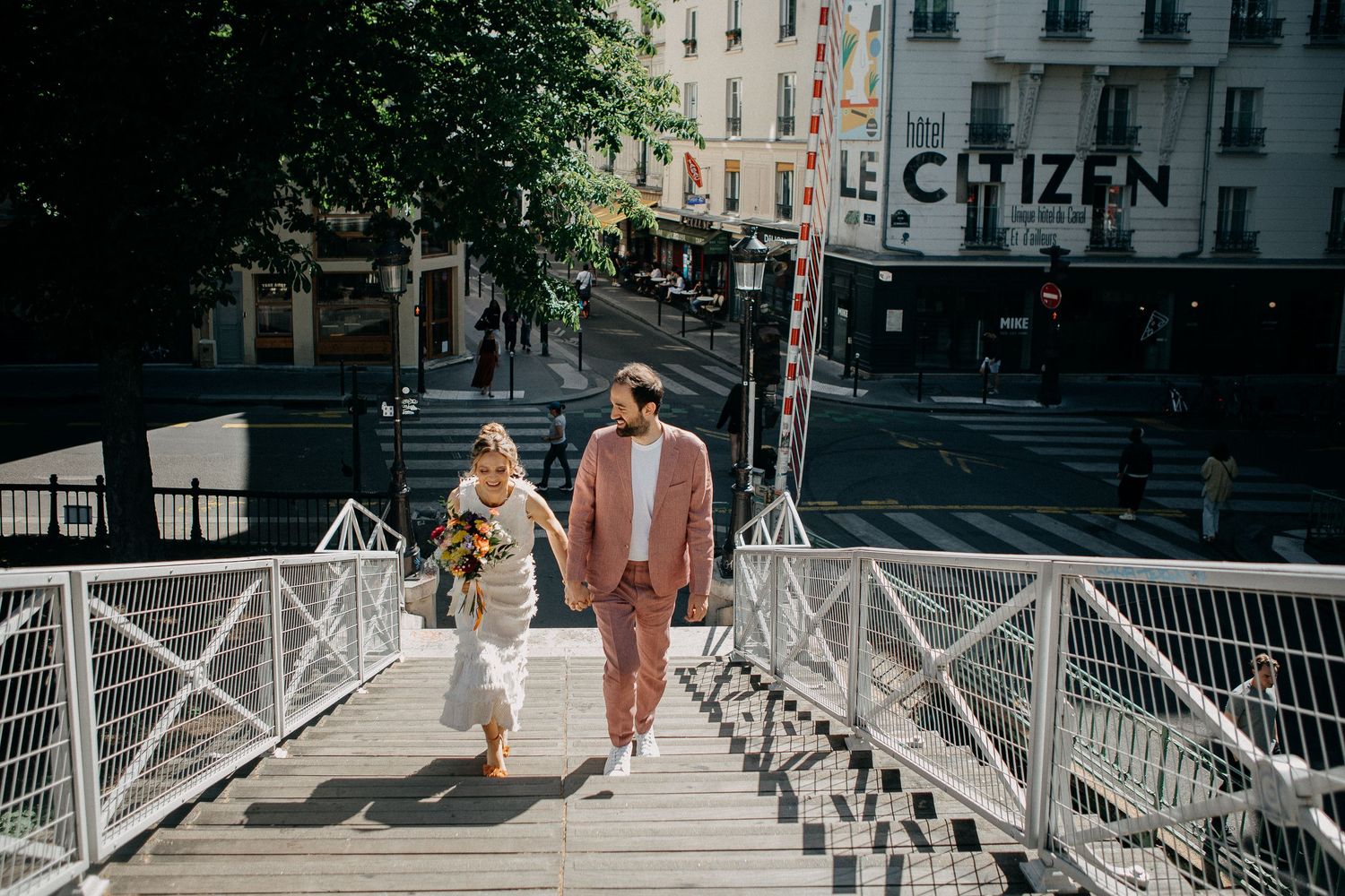 Couple holding hands walking on a bridge in Paris with Le Citizen hotel in the background on a sunny day.