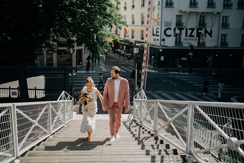 Couple holding hands walking on a bridge in Paris with Le Citizen hotel in the background on a sunny day.