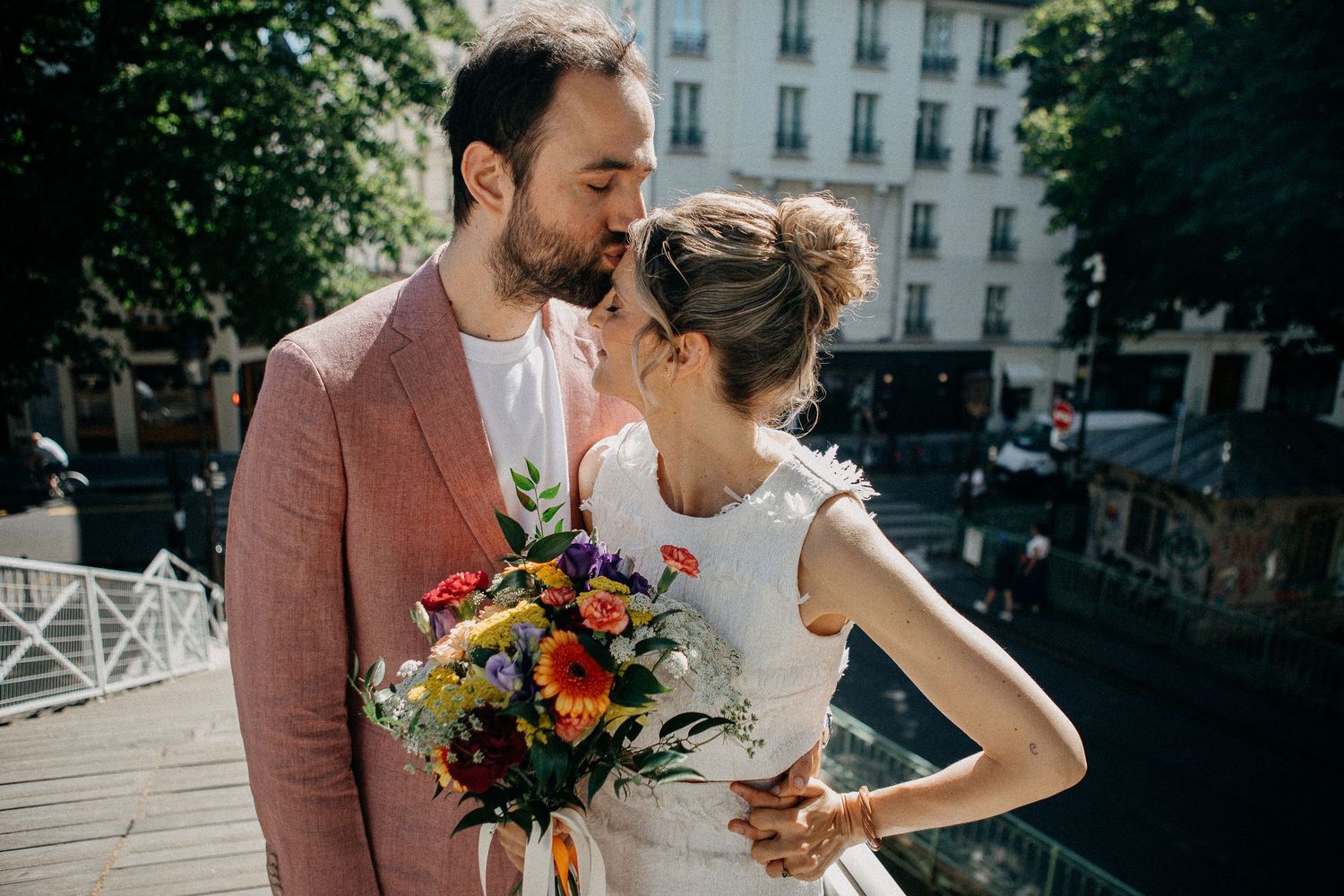 Couple sharing a tender kiss on a bridge in Paris, bride holding a vibrant colorful bouquet.