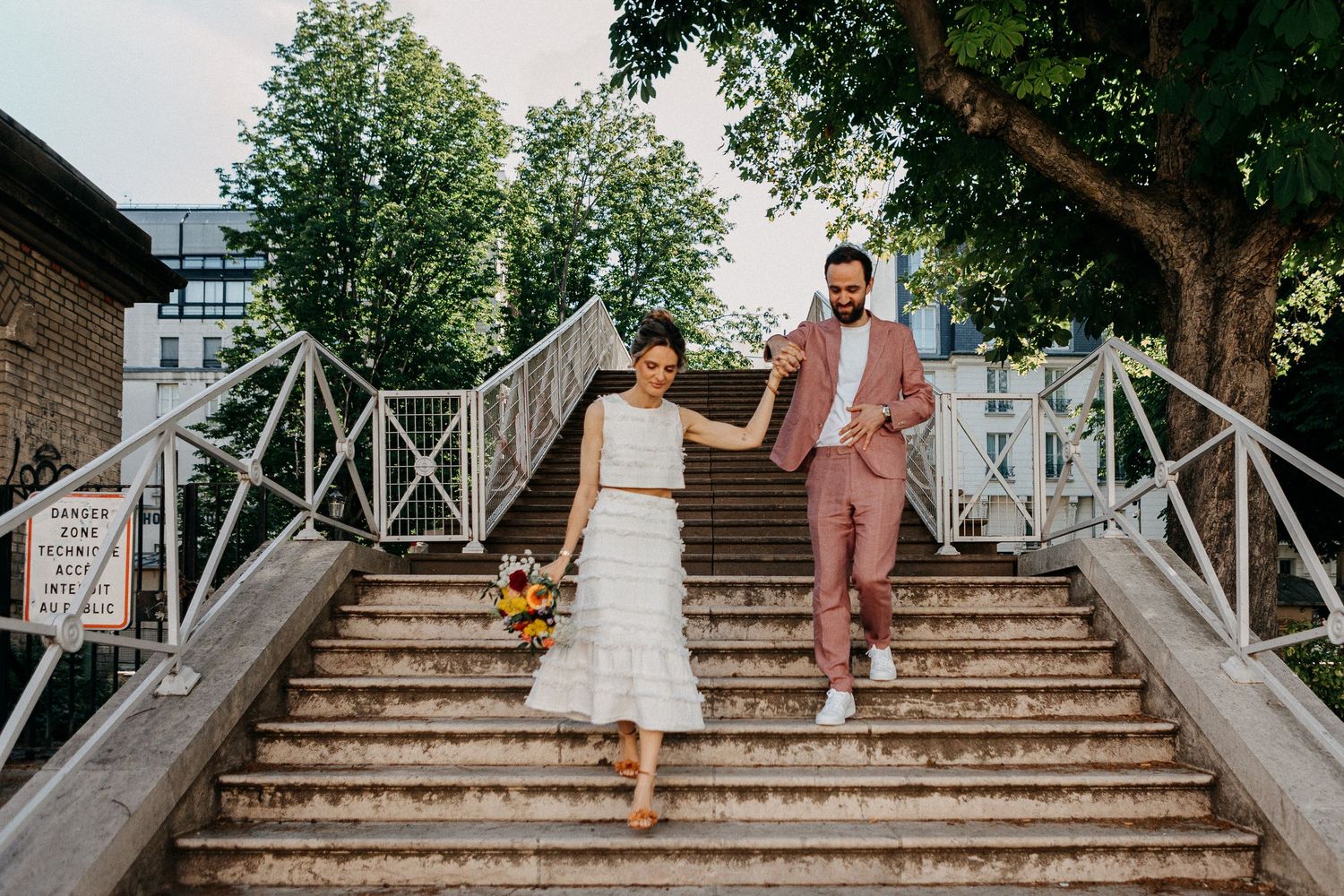 Bride and groom holding hands walking down outdoor stone steps, bride in white dress holding bouquet, groom in pink suit.
