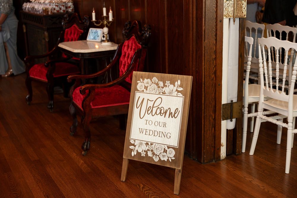 Elegant wedding welcome sign on wooden easel with floral design, red velvet chairs and candlelit table in background.