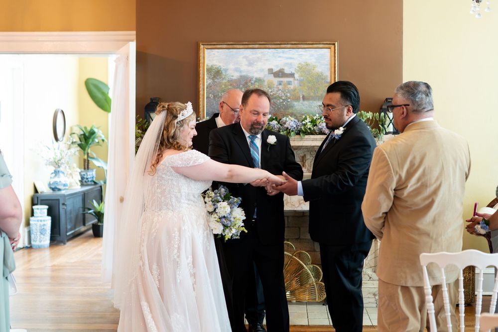 Bride in white gown greets groom during indoor wedding ceremony near floral fireplace.