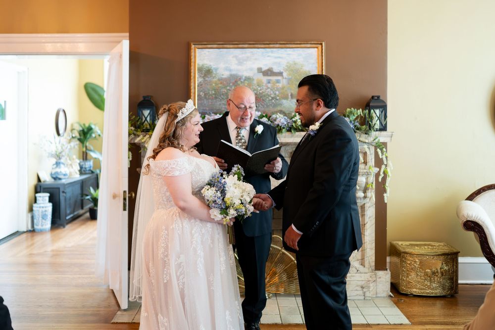 Bride and groom exchange vows with officiant at intimate indoor wedding ceremony near fireplace.