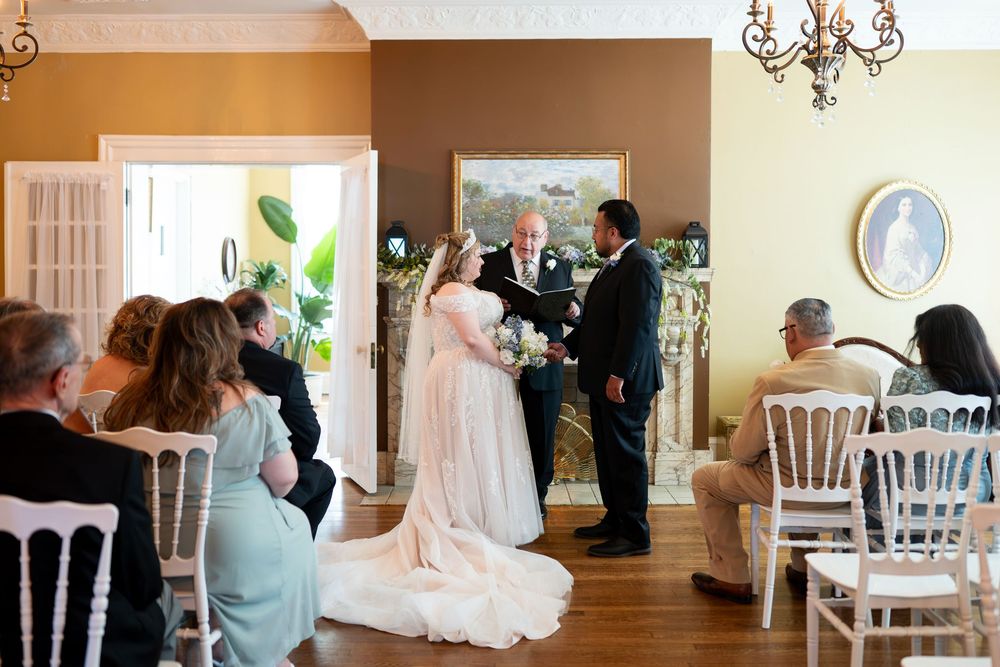 Elegant indoor wedding ceremony with bride in white gown, groom, and officiant before ornate fireplace mantel.