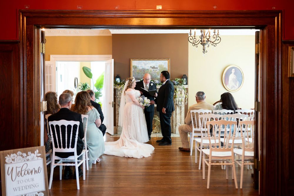 Elegant indoor wedding ceremony with bride and groom at the altar, guests seated in white chairs in a warmly decorated venue.