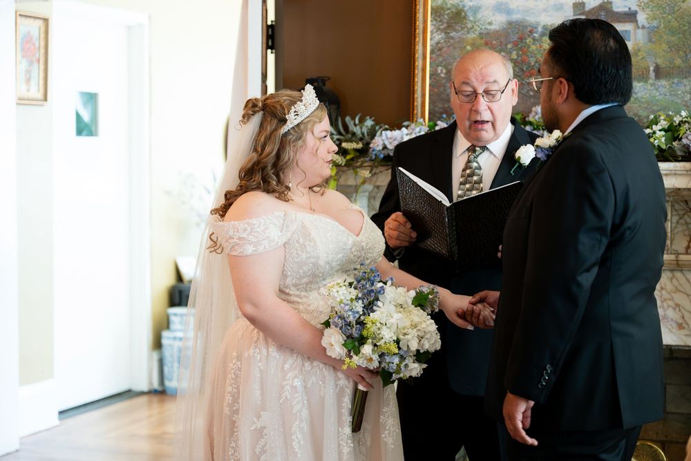 Bride in lace gown holding bouquet exchanges vows with groom before officiant during intimate indoor wedding ceremony.