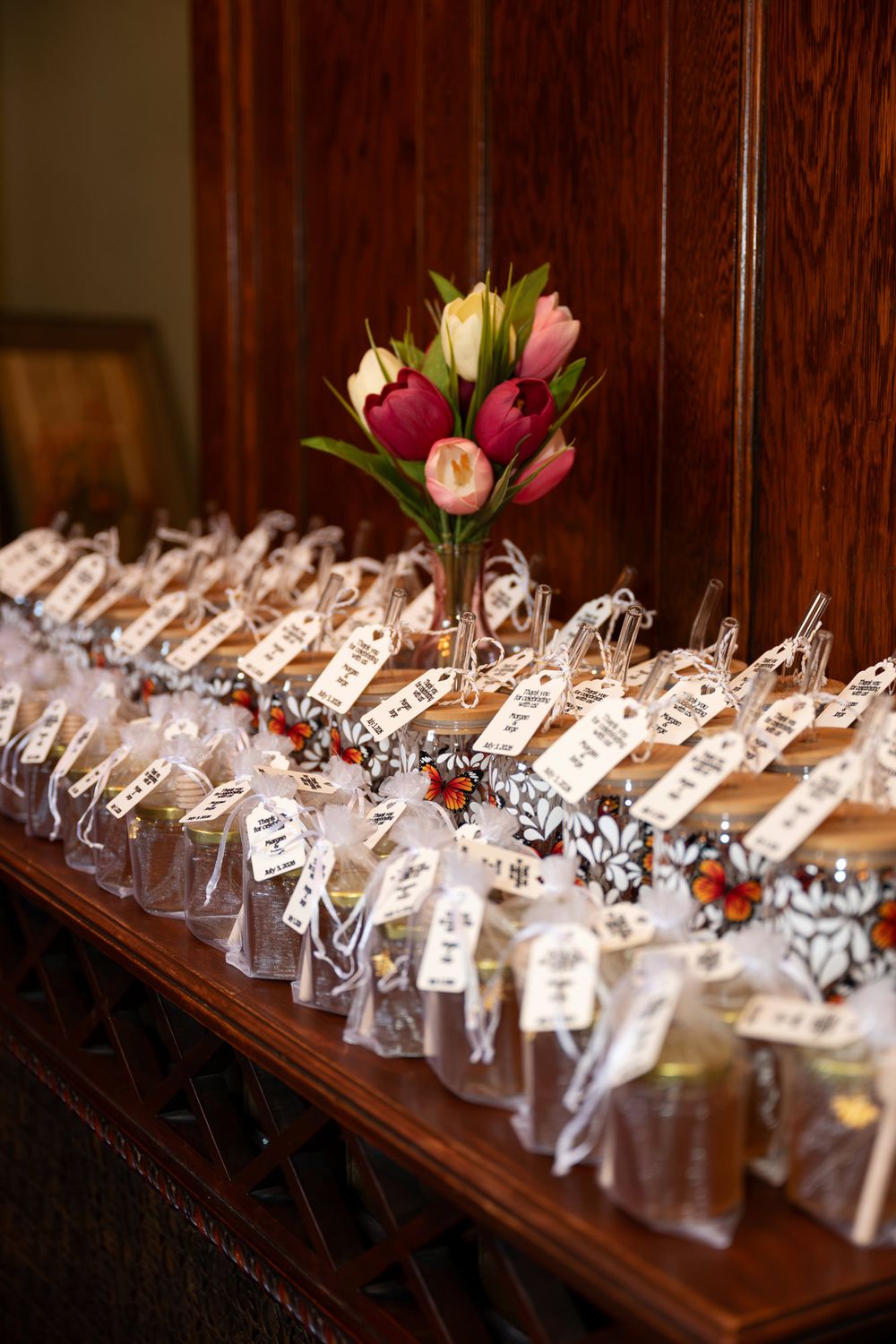 Wedding favor jars with tags displayed on wooden table beside pink and red tulip floral arrangement.