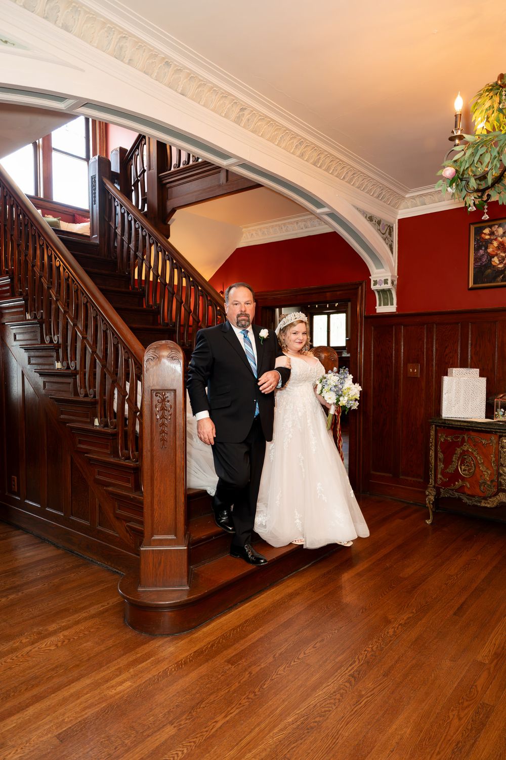 Bride in white gown holds bouquet while escorted by suited man down elegant wooden staircase in ornate venue.