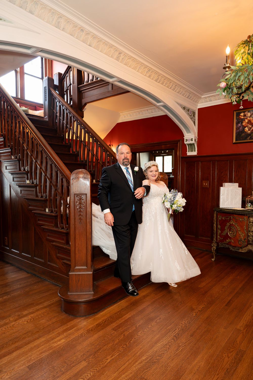 Bride and groom descend elegant wooden staircase in historic venue, bride holding white bouquet.