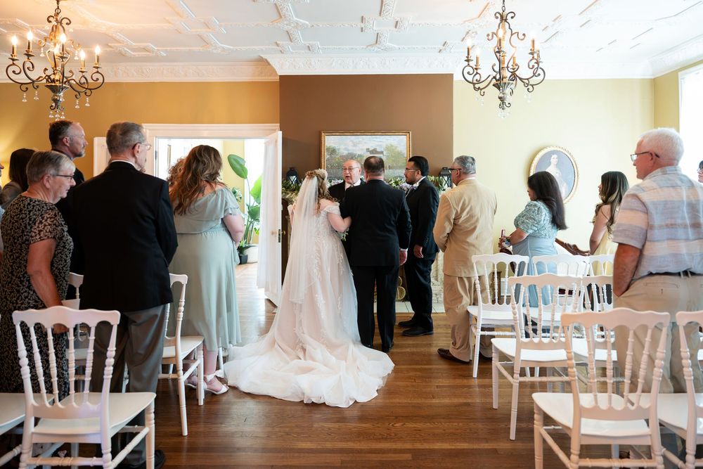 Indoor wedding ceremony with bride in white gown, guests seated in white chairs, elegant chandelier venue.