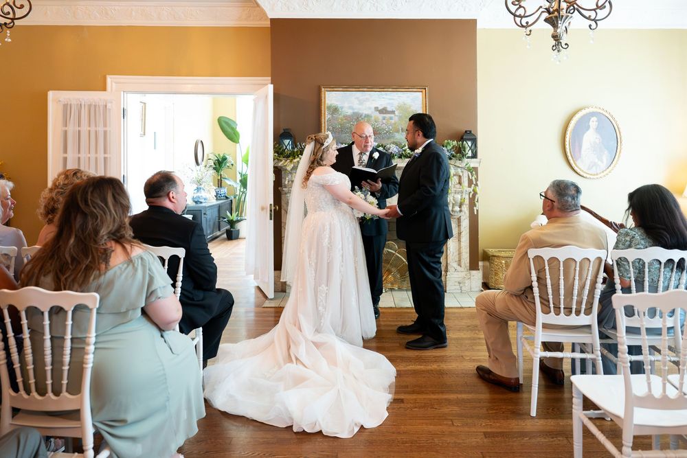 Elegant indoor wedding ceremony with bride and groom at altar, guests seated in white chairs in a warmly decorated venue.