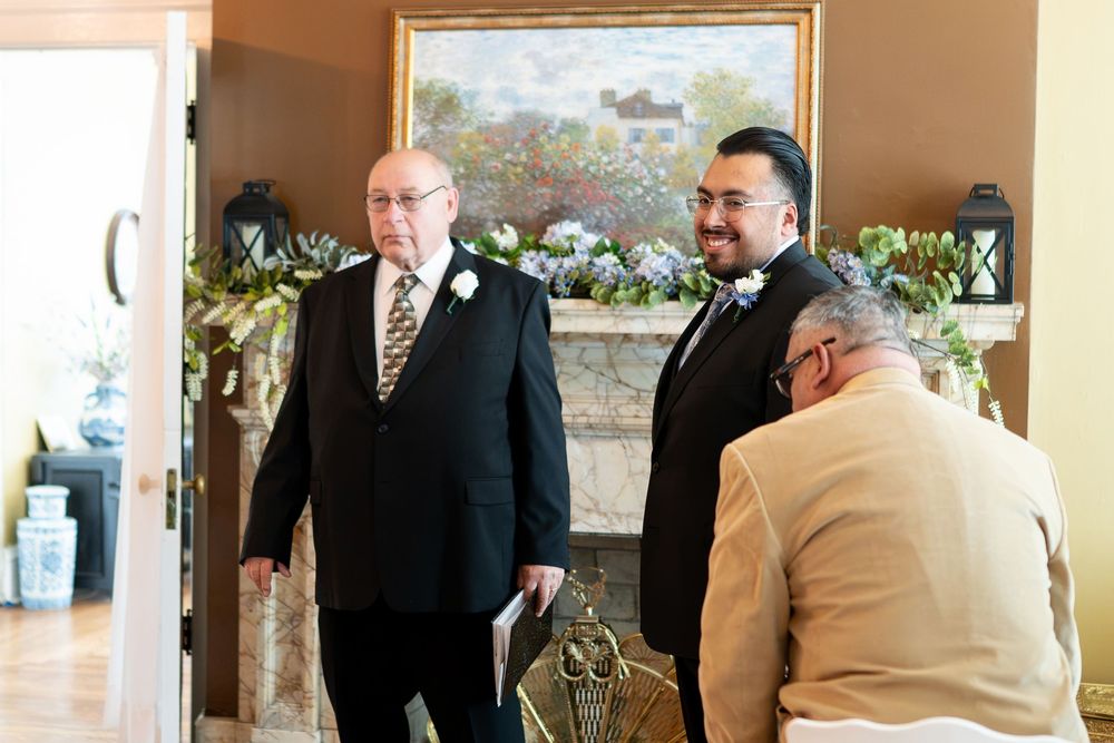 Two men in formal attire share a moment before a wedding ceremony near a decorated fireplace.