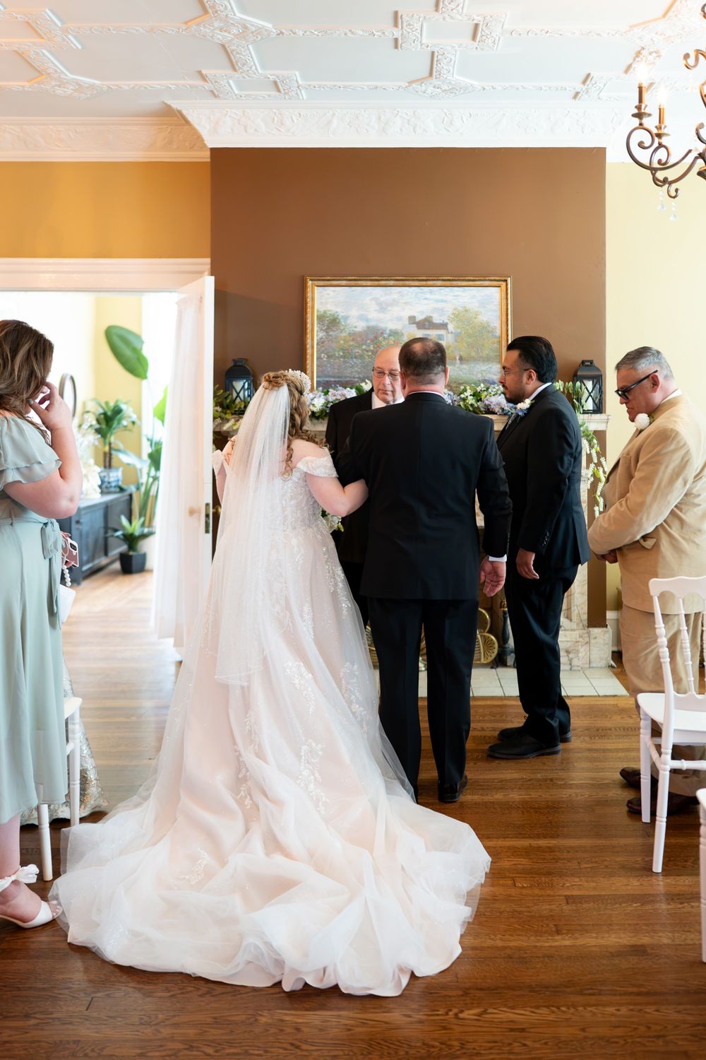 Bride in lace gown with cathedral veil stands at indoor wedding ceremony with groom and officiant.