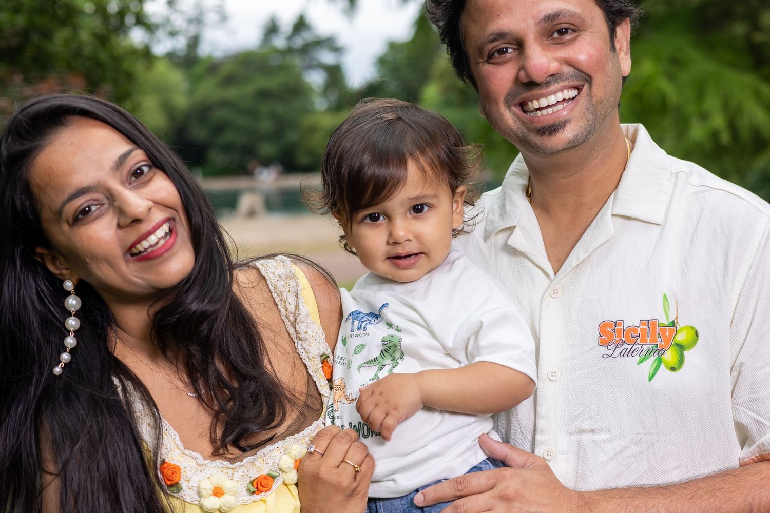 A smiling South Asian family with a mother, father, and baby posing outdoors in a lush green park setting.