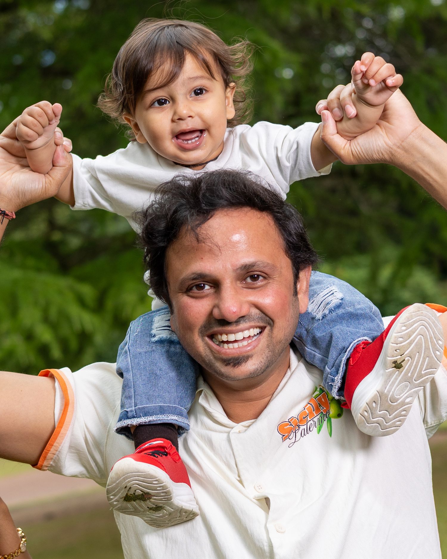 A smiling man carries a happy toddler on his shoulders outdoors, both wearing white shirts.