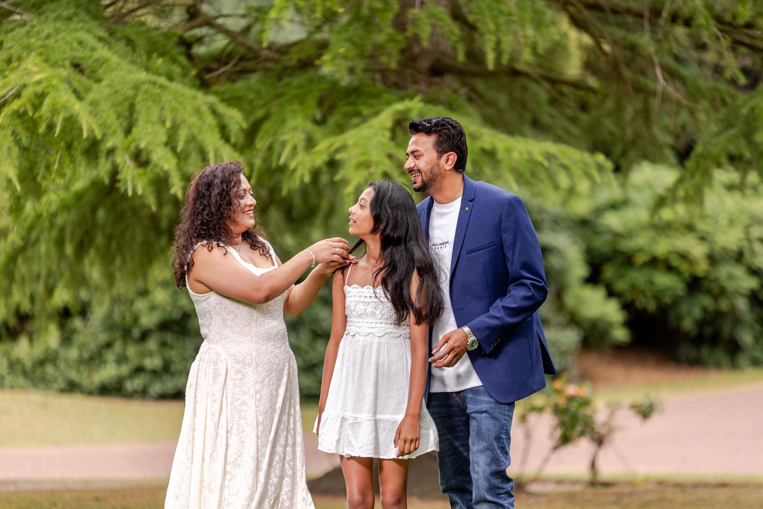A joyful South Asian family of three smiling outdoors in a lush green park, wearing elegant white and blue outfits.