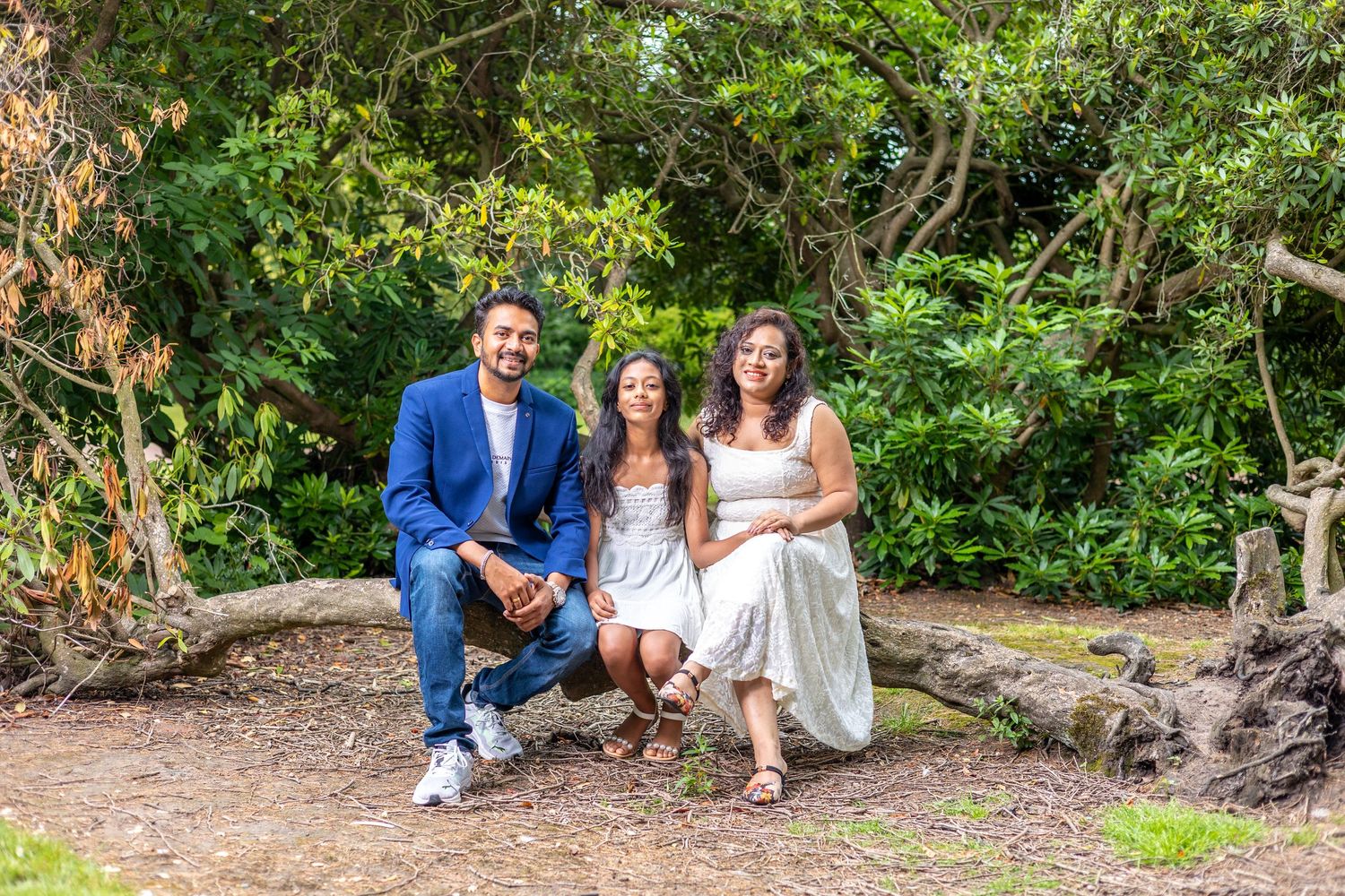 Family of three posing outdoors on a log surrounded by lush green trees in a beautiful garden setting.