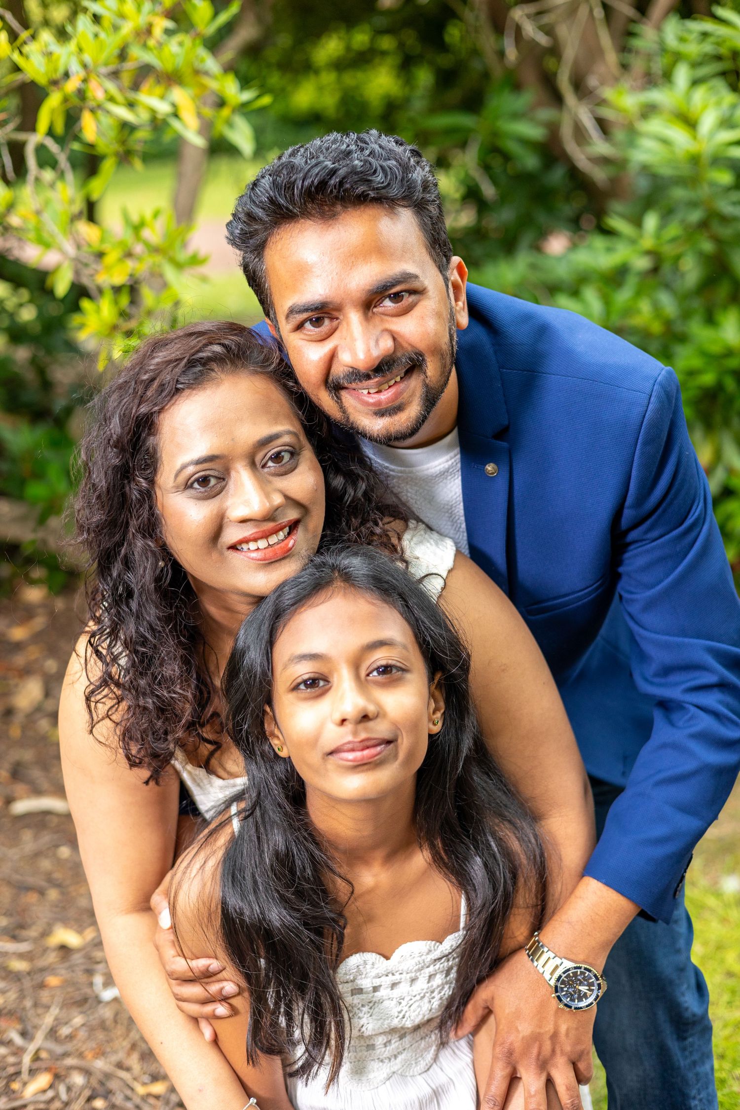 A smiling South Asian family of three poses outdoors, with parents embracing their young daughter in a lush green garden.
