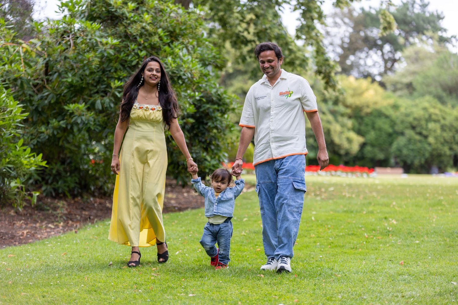 A happy family of three walks hand-in-hand through a lush green park, parents swinging their toddler between them.