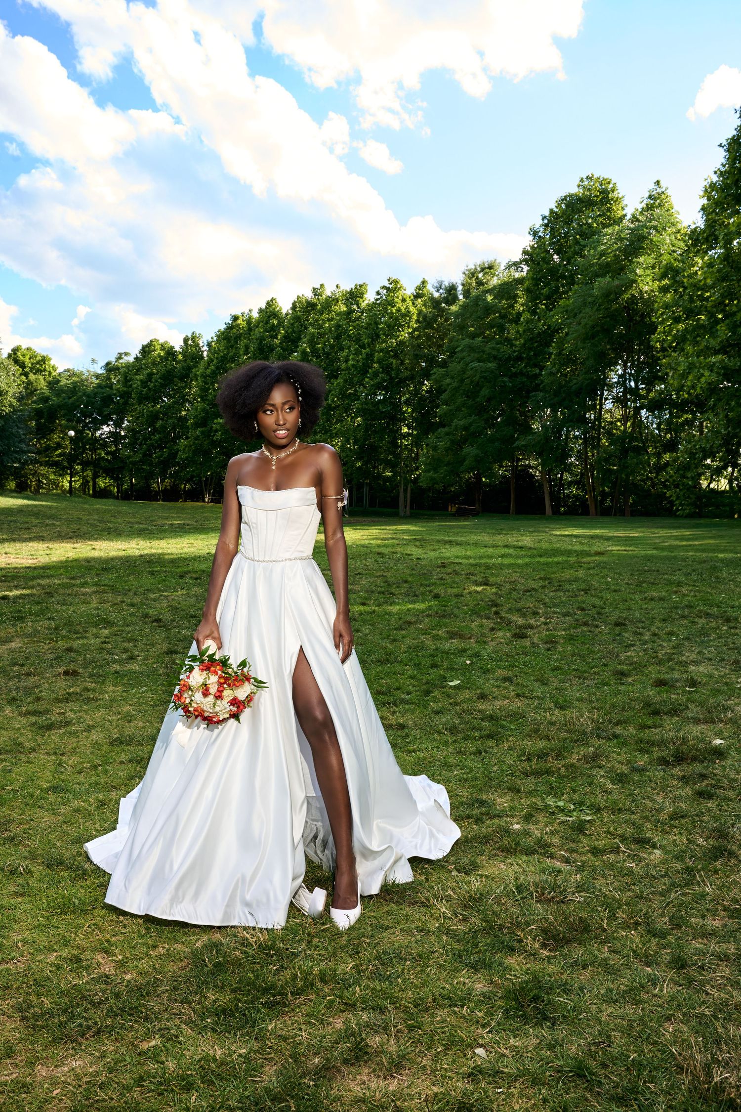 A beautiful Black bride in a flowing white gown holds a colorful bouquet while gazing upward in a lush green garden.