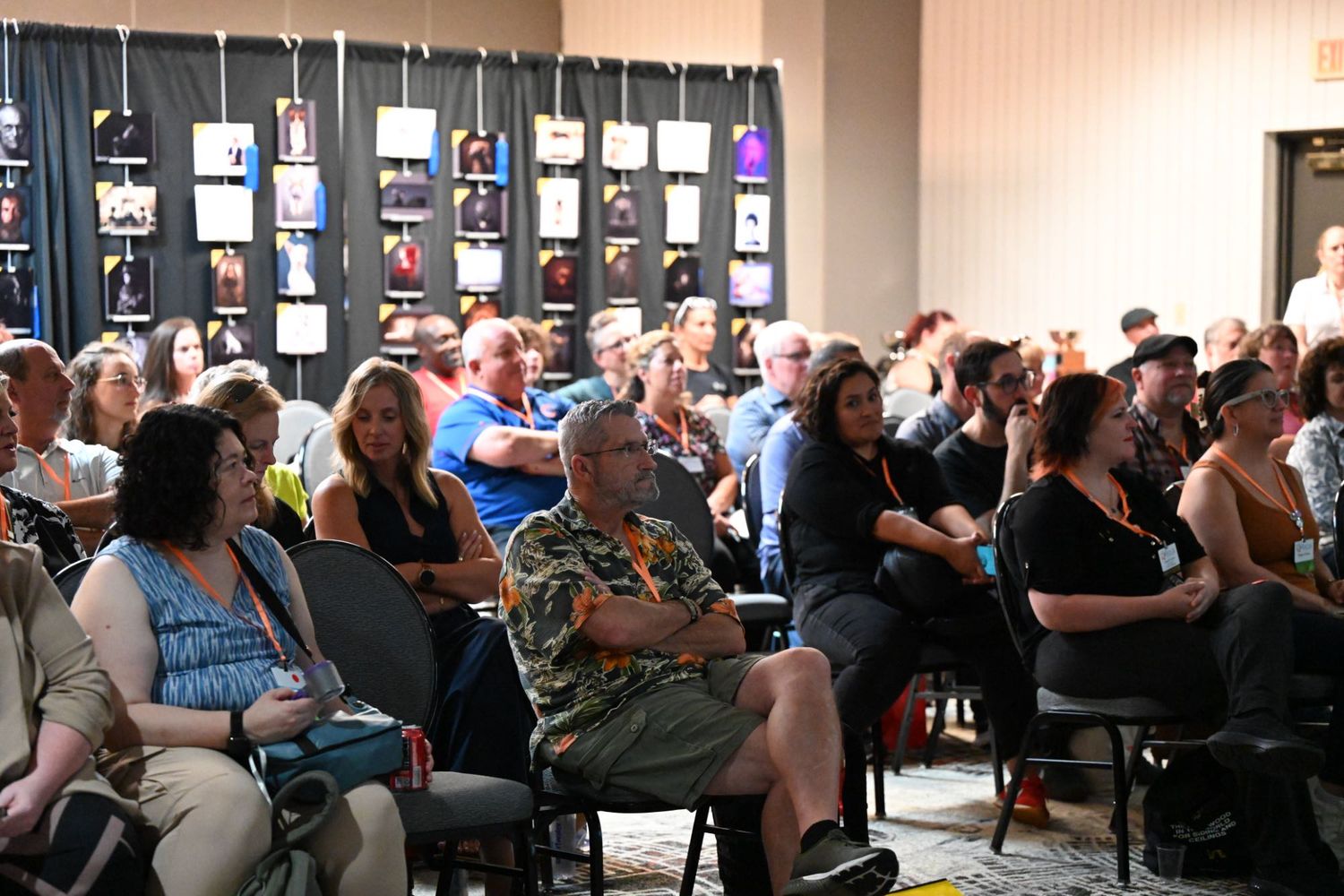 A large audience seated in chairs at an indoor event with colorful photo displays on the wall behind them.