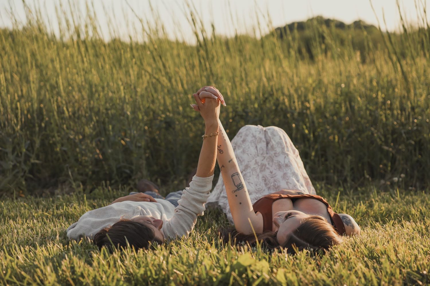 Two women lie in tall grass with legs raised, enjoying a sunny outdoor moment together.
