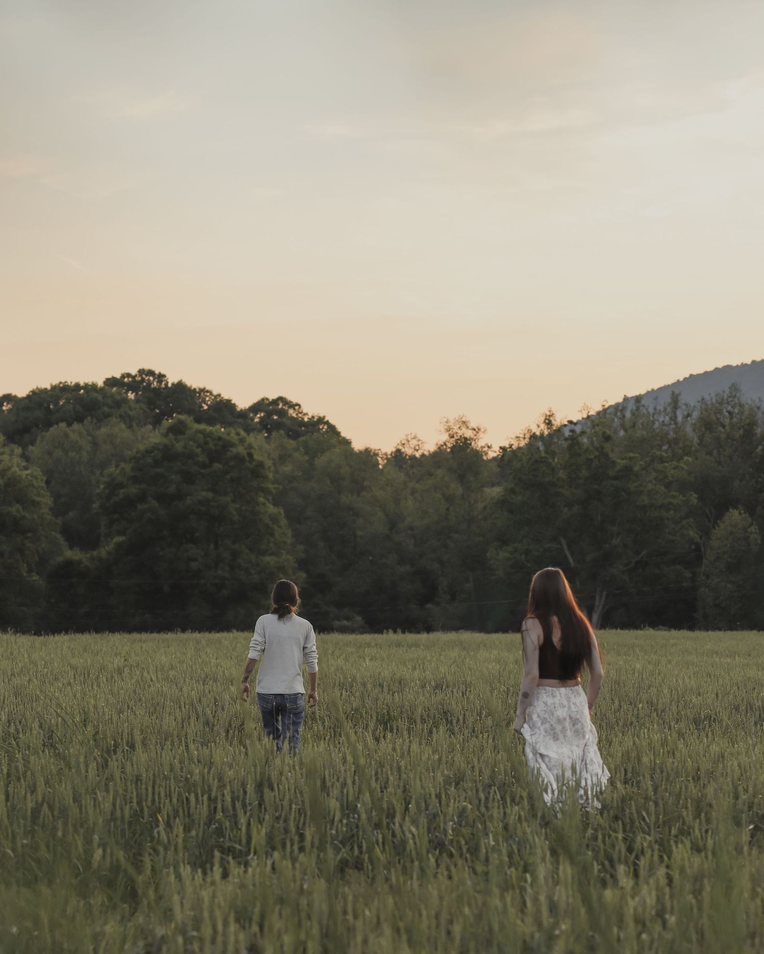 Two women walk apart in a vast green field at dusk with trees and a mountain behind.