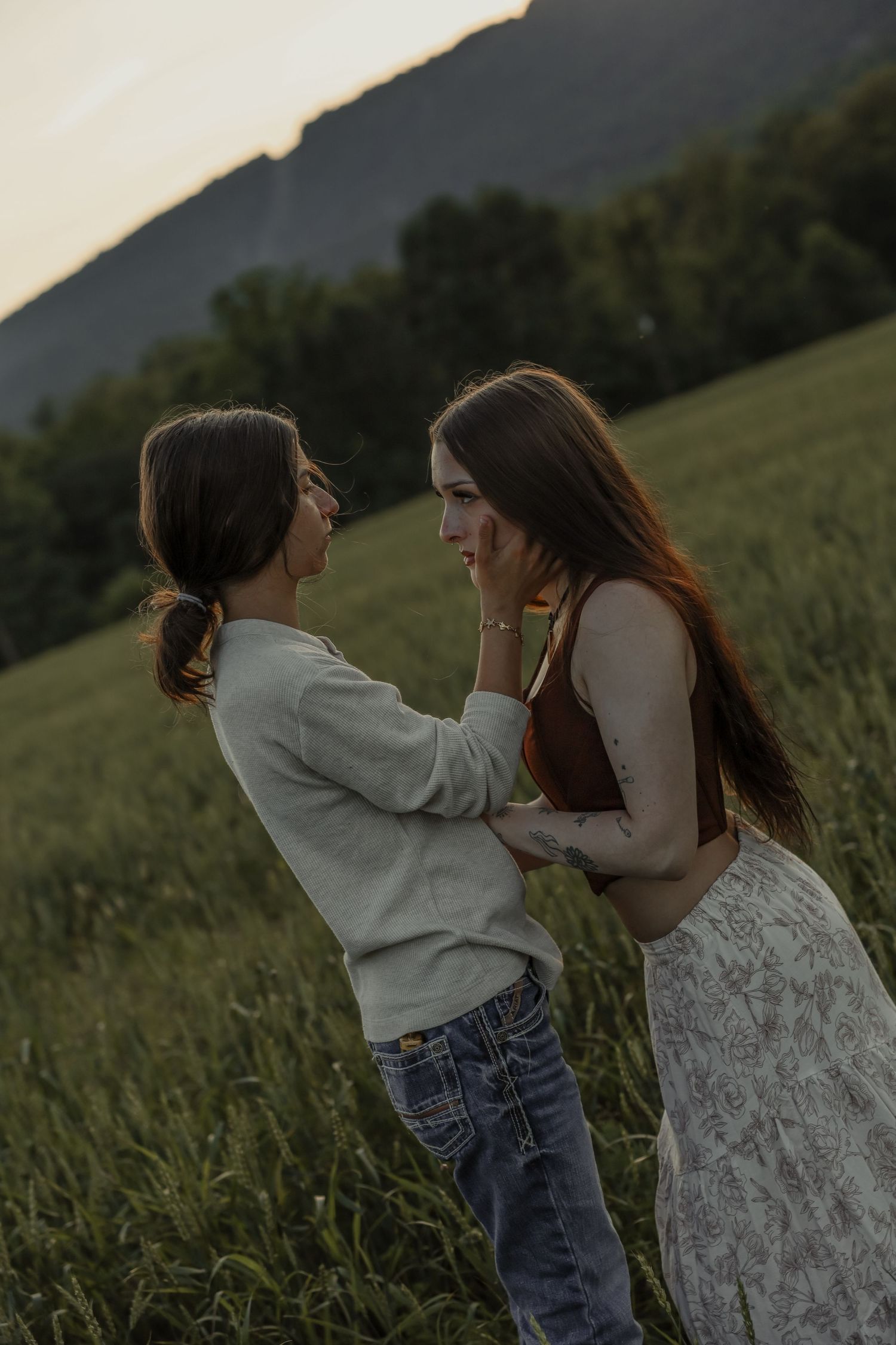 Two women face each other intimately in a green field at dusk, one touching the other's face gently.
