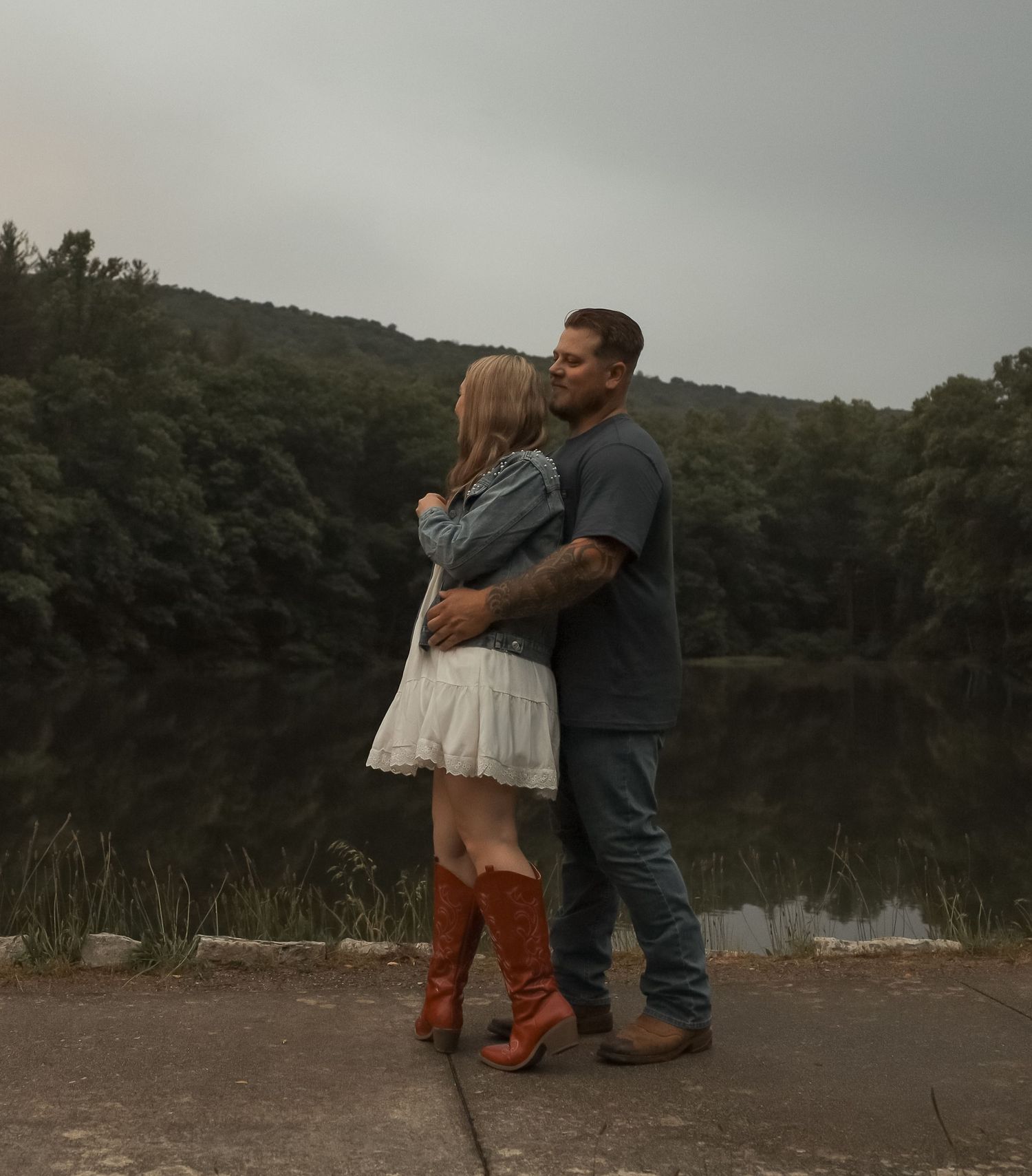 Romantic couple kissing near a serene lake with green hills in the background on overcast day.