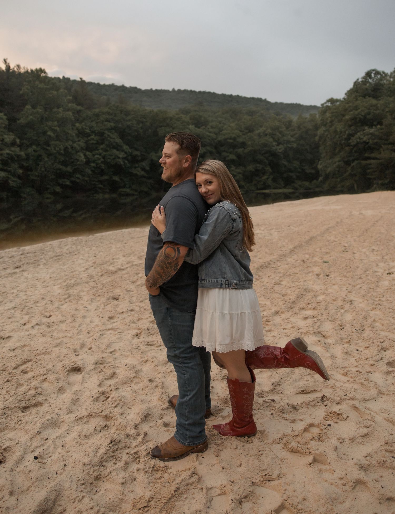 Couple embracing on a sandy riverbank with green forested hills in the background.