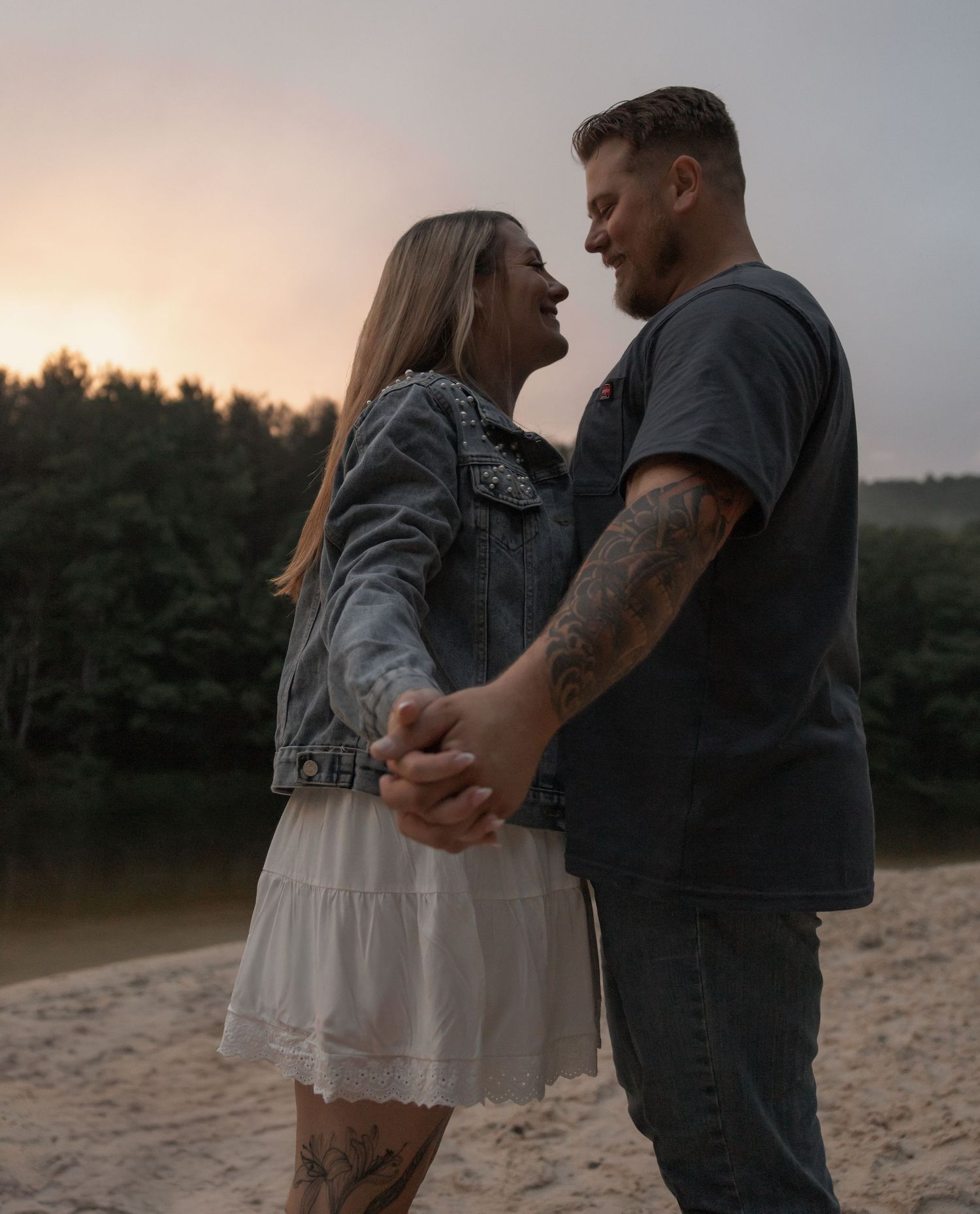 A couple holds hands and smiles at each other on a sandy beach at sunset, woman in white skirt and denim jacket.