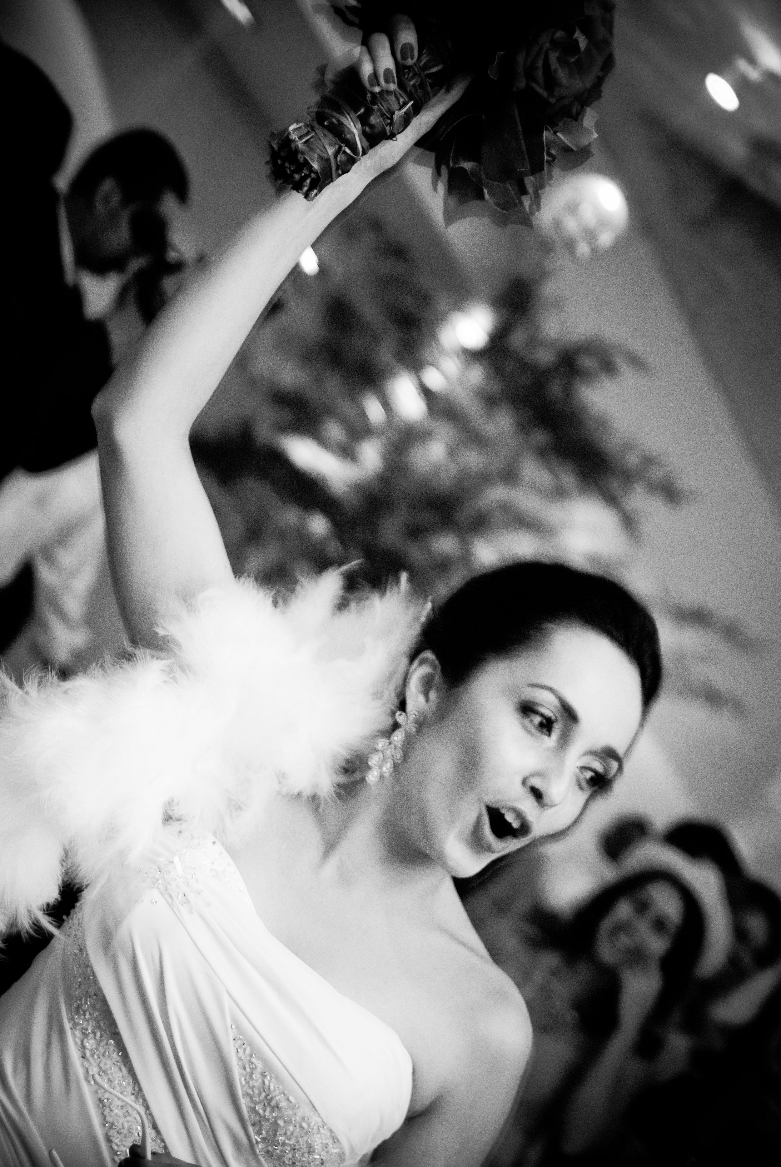 Black and white photo of an expressive bride in a white feather boa raising a bouquet at her reception.