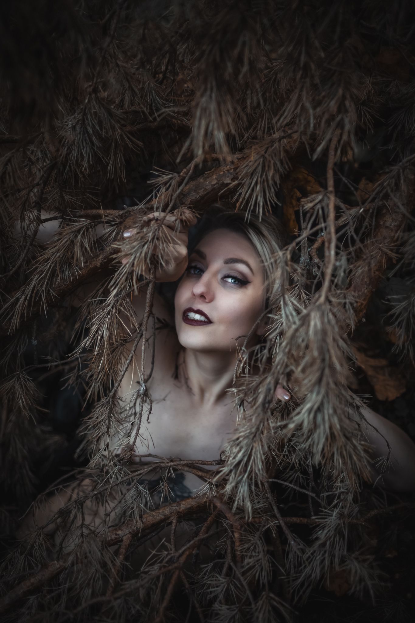 A mysterious blonde woman with dark makeup peers through dried pine branches in a moody, dark forest setting.