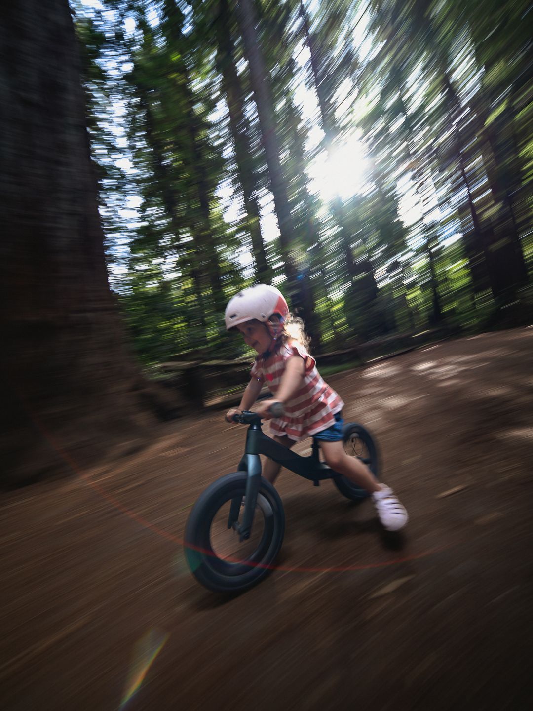 Young girl in pink helmet rides a balance bike through a sunlit forest trail, motion blur capturing her speed.