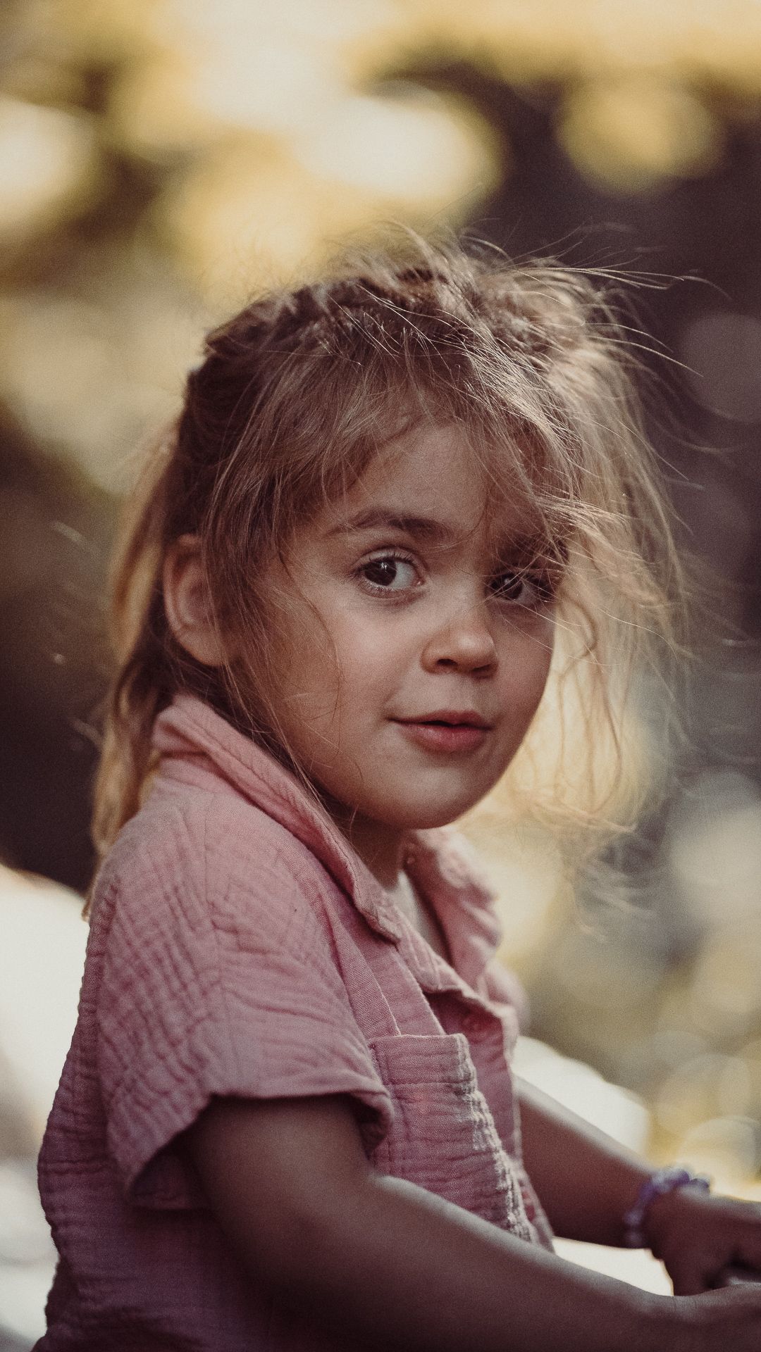 Young girl with messy hair in pink shirt gazes at camera outdoors in soft bokeh light.
