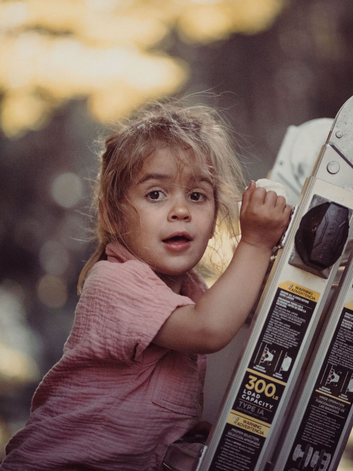 A young girl with wide eyes clings to a ladder outdoors, wearing a pink shirt, with a blurred bokeh background.
