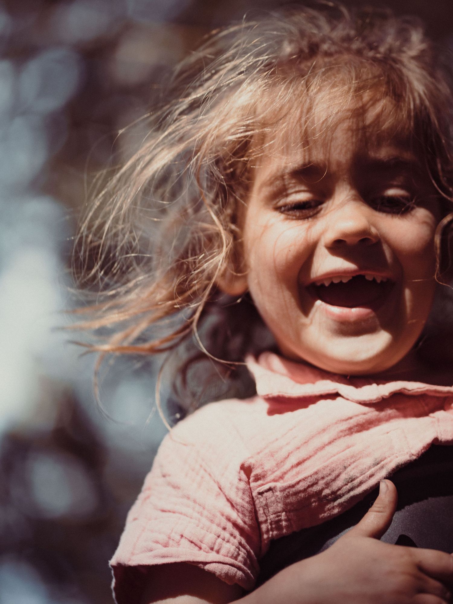 A joyful young girl with windswept hair smiles broadly outdoors in warm sunlight, wearing a pink shirt.