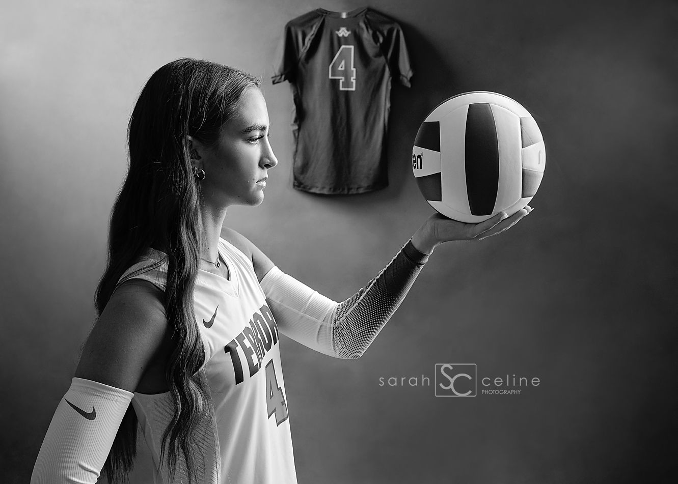Black and white photo of a female volleyball player holding a ball, with jersey number 4 hanging on wall behind her.
