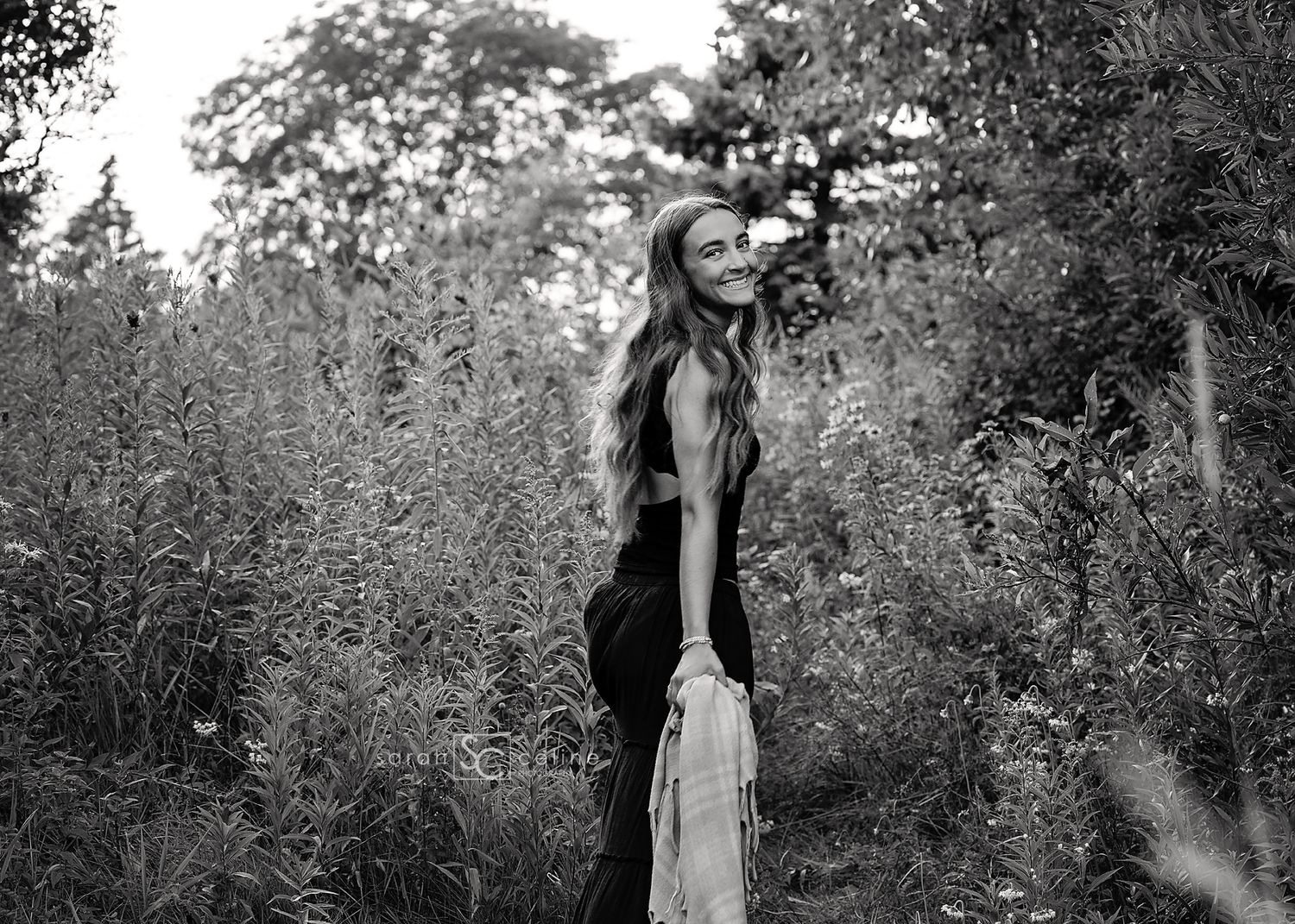Black and white photo of a young woman with long hair standing among tall plants in a lush outdoor setting.