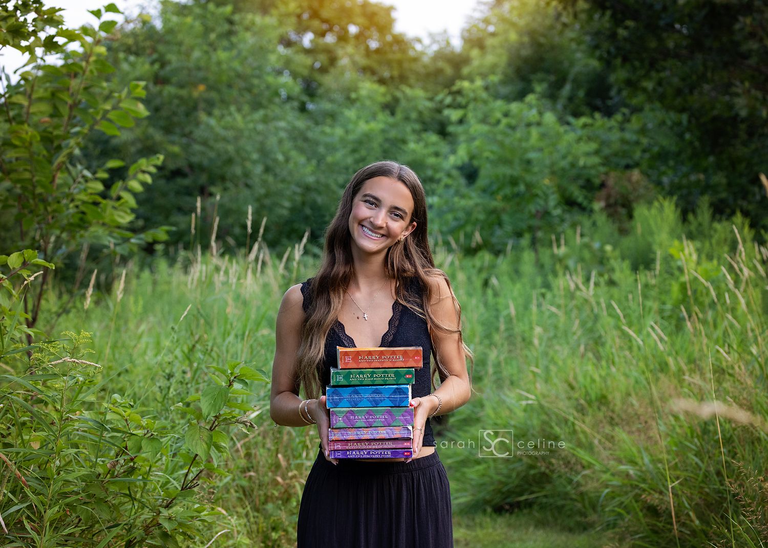 Smiling teenage girl holding colorful stacked books outdoors in a lush green meadow setting.