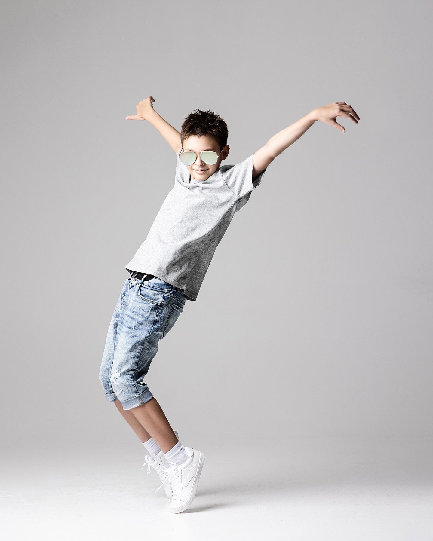 A young boy in jeans, white tee, and sunglasses strikes a dynamic dance pose with arms outstretched.